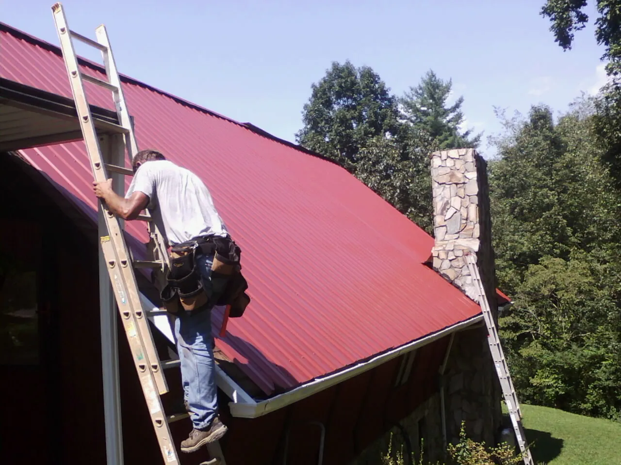 Roofer on a ladder beside a red metal roof and stone chimney — Soffit & Fascia Repair by Roofing Brothers in Ocean Grove